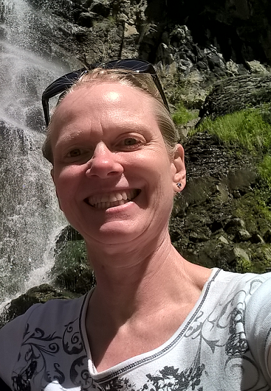 A woman smiling as she takes a selfie in front of a majestic waterfall, capturing a moment of joy and adventure.